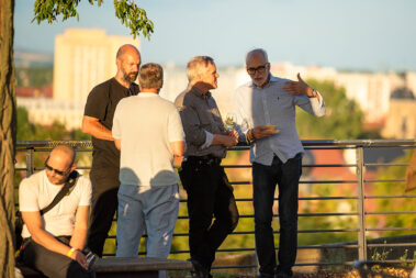 Vier Männer stehen an einem Geländer, unterhalten sich, essen und trinken dabei