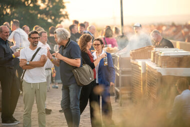 Menschen unterhalten sich, haben ein Getränk in der Hand, auf der rechten Seite befindet sich die Buffetstrecke